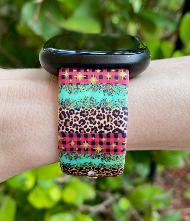Op de afbeelding: Een zwarte smartwatch met een kleurrijk kerstbandje. Het bandje heeft een rood-zwart ruitpatroon, groene dennentakken, gouden sterren en een luipaardprint.