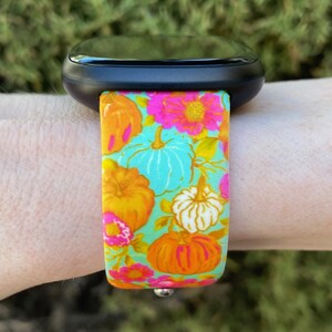 May include: A black smartwatch with a colorful floral and pumpkin print band. The band features a turquoise background with orange, pink, and white pumpkins and flowers.