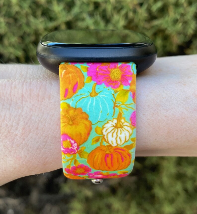 May include: A black smartwatch with a colorful floral and pumpkin print band. The band features a turquoise background with orange, pink, and white pumpkins and flowers.