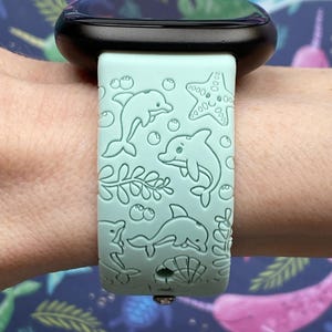 May include: A light blue silicone watch band with a dolphin and starfish design. The band is on a black smartwatch.