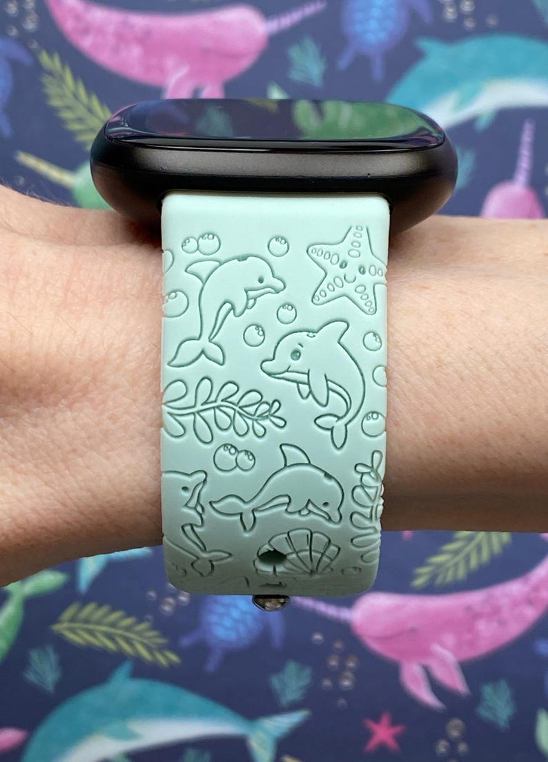 May include: A light blue silicone watch band with a dolphin and starfish design. The band is on a black smartwatch.