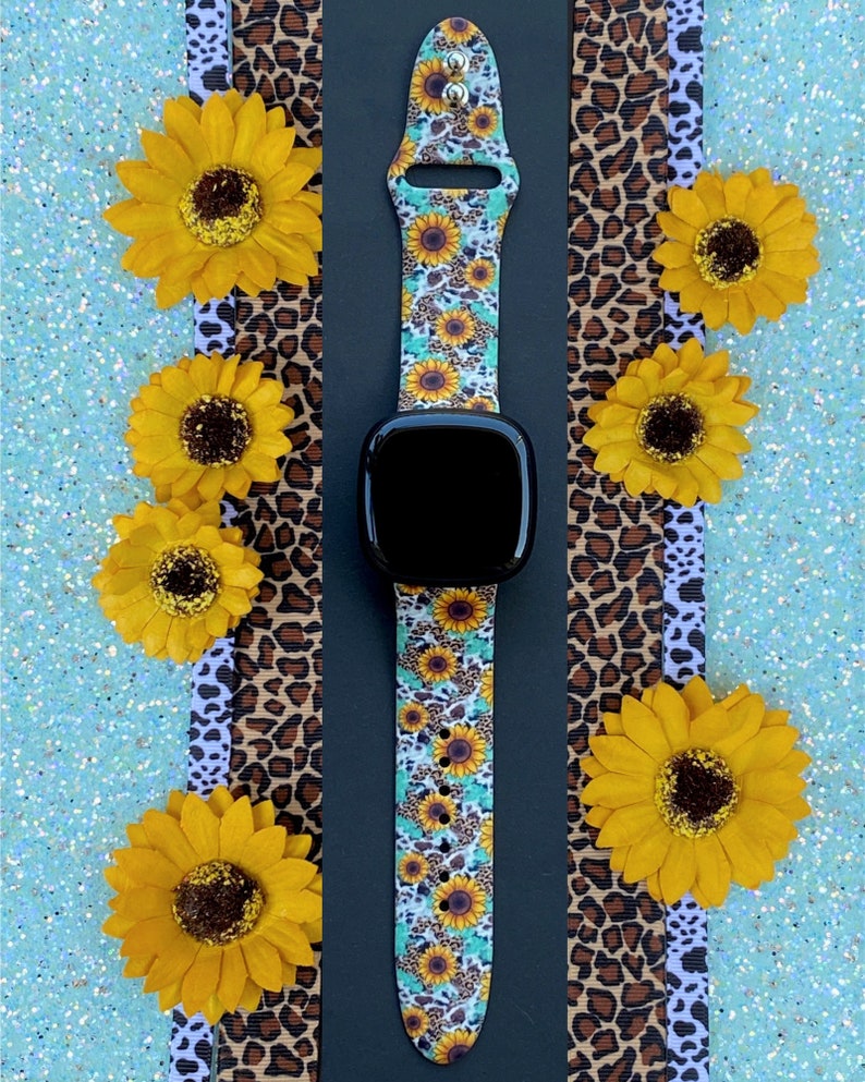May include: A black smartwatch with a floral and leopard print band. The band features yellow sunflowers on a light blue and white background. The watch is surrounded by yellow sunflowers and leopard print ribbons.