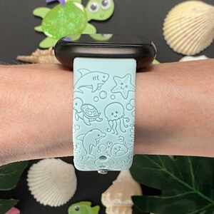 May include: A light blue silicone watch band with a sea-themed design. The band features a shark, starfish, turtle, jellyfish, dolphin, and crab.