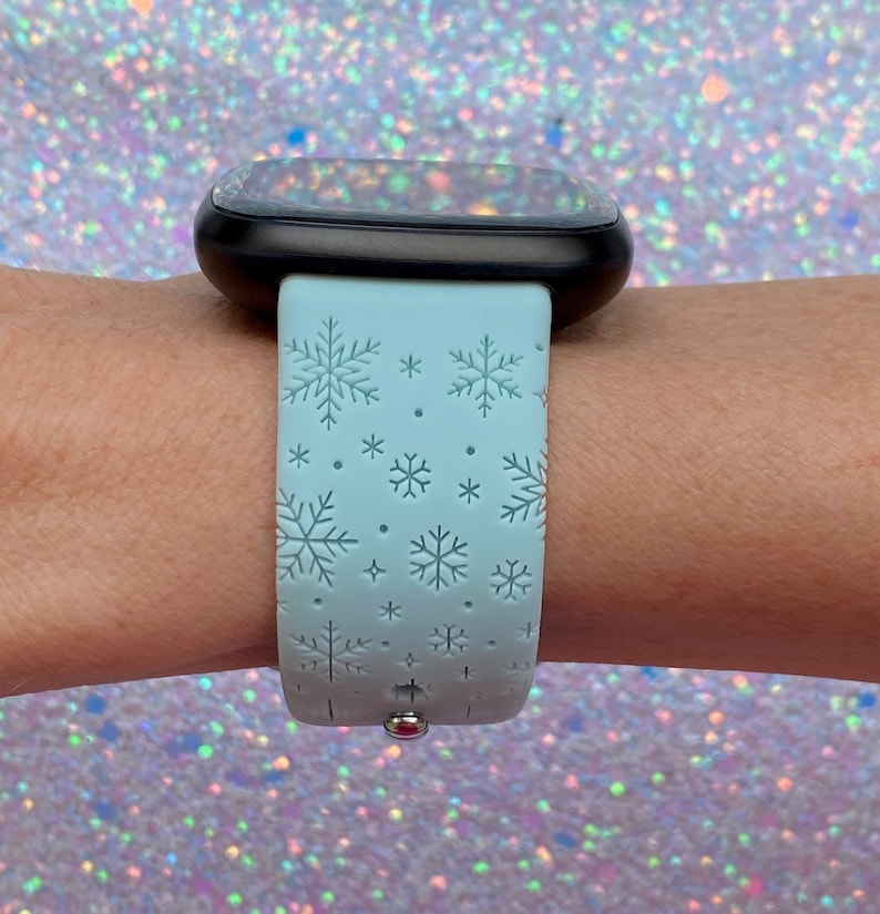 May include: A light blue smartwatch band with a snowflake pattern. The band is made of silicone and has a silver buckle.