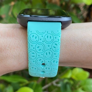 May include: A turquoise smartwatch band with a smiley face pattern. The band is made of silicone and has a black watch face.
