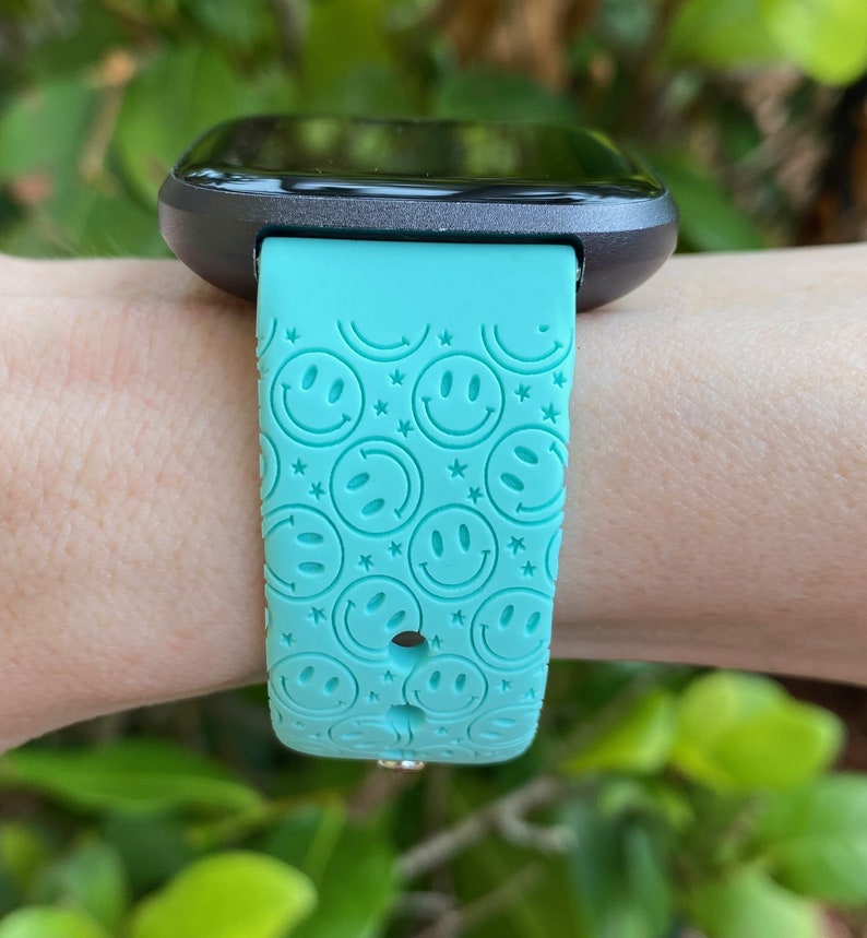 May include: A turquoise smartwatch band with a smiley face pattern. The band is made of silicone and has a black watch face.
