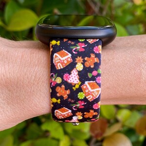 May include: A black smartwatch with a festive band featuring gingerbread houses, gingerbread men, candy canes, and Christmas trees on a black background. The band is attached to a black watch face.