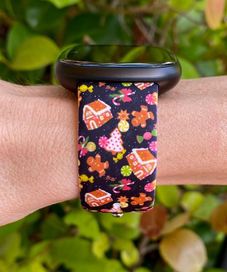 May include: A black smartwatch with a festive band featuring gingerbread houses, gingerbread men, candy canes, and Christmas trees on a black background. The band is attached to a black watch face.