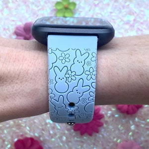 K&ouml;nnte beinhalten: Hellblaues Uhrenarmband mit einem sich wiederholenden Muster aus Hasen- und Blumendesigns. Das Armband ist an einer schwarzen Smartwatch befestigt. Das Armband besteht aus einem weichen, flexiblen Material.