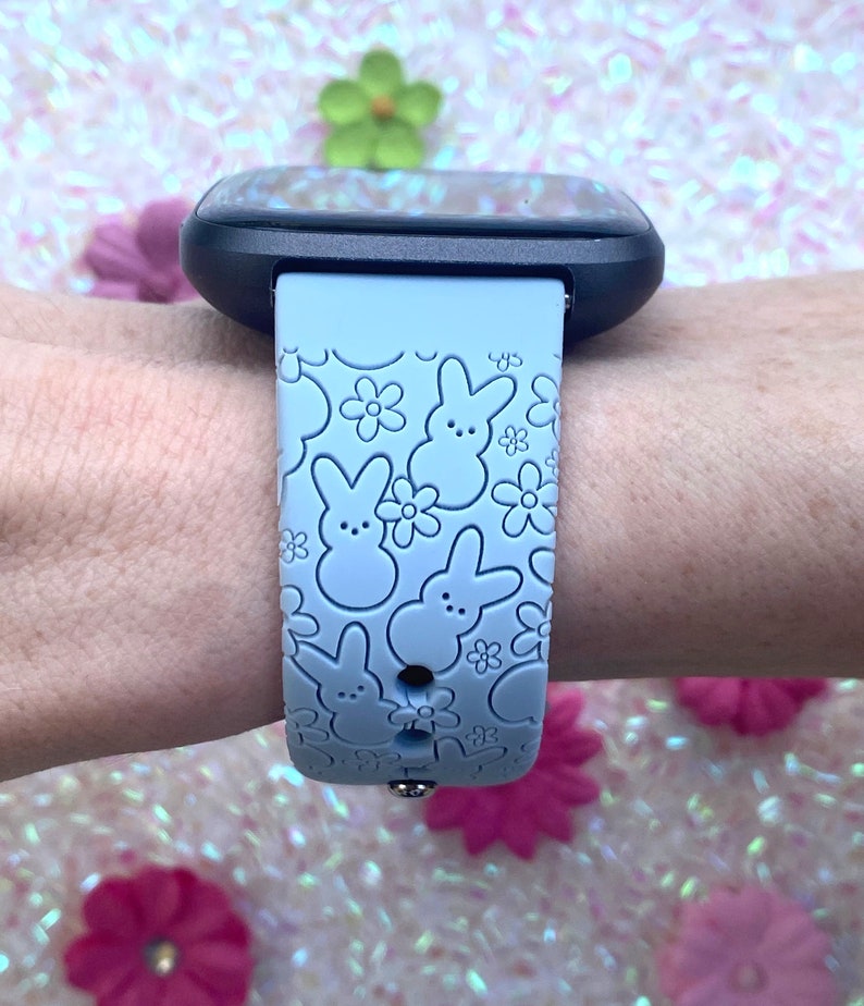 K&ouml;nnte beinhalten: Hellblaues Uhrenarmband mit einem sich wiederholenden Muster aus Hasen- und Blumendesigns. Das Armband ist an einer schwarzen Smartwatch befestigt. Das Armband besteht aus einem weichen, flexiblen Material.