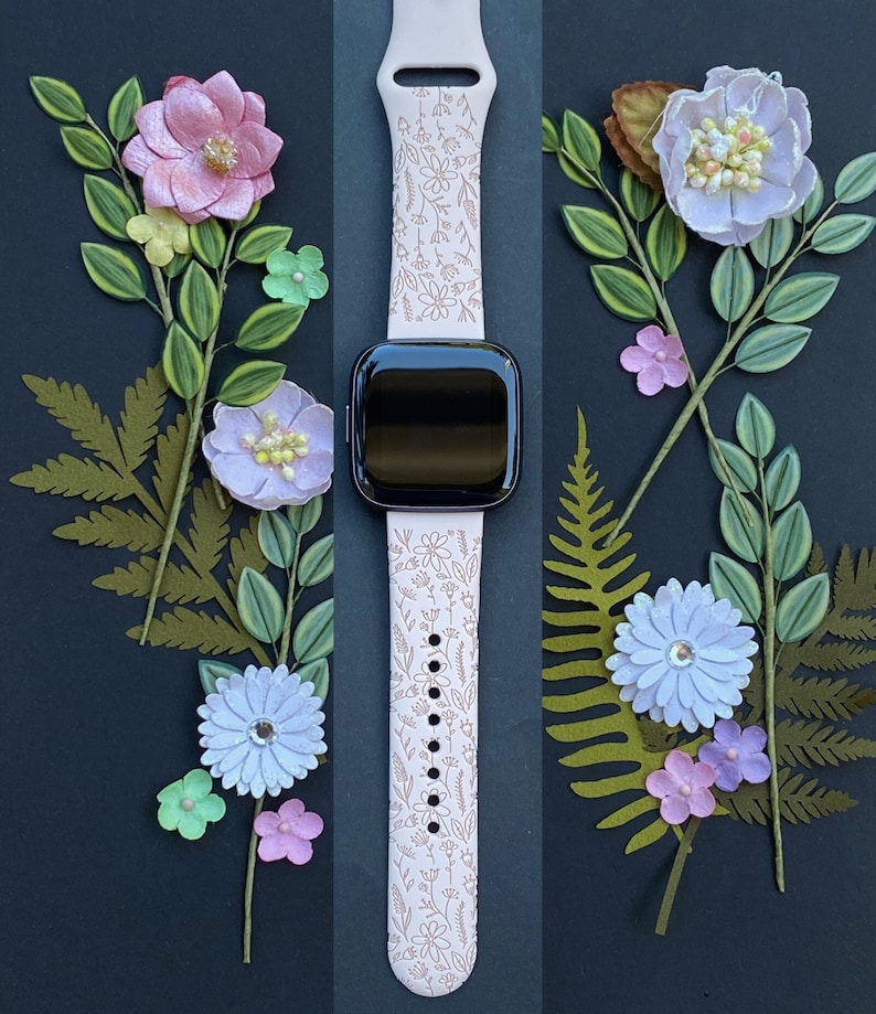 Op de afbeelding: Een roze horlogebandje met een bloemenpatroon. Het bandje is gemaakt van siliconen en heeft een zwart horlogeglas. Het bandje is omringd door papieren bloemen in verschillende kleuren.