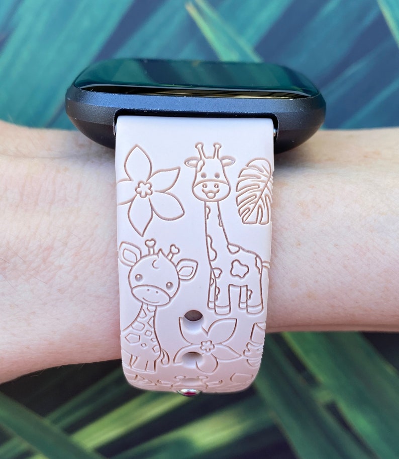 May include: A pink silicone watch band with a giraffe and flower design. The band is compatible with a Fitbit watch.