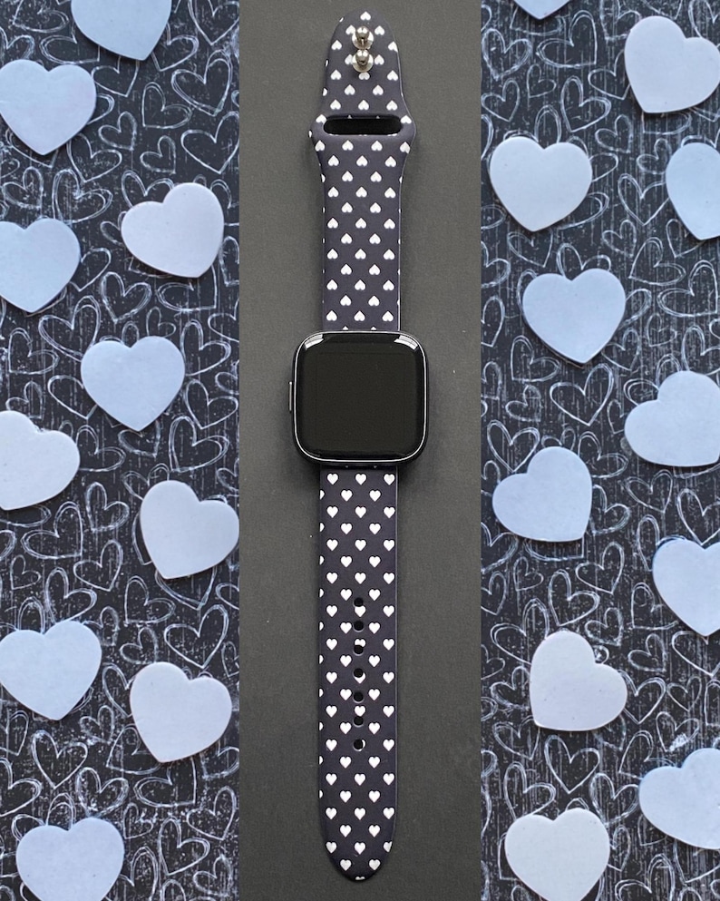 May include: A smartwatch with a black screen and a navy blue band patterned with small white hearts. The watch is centered against a dark background with light blue heart-shaped cutouts and a heart-themed pattern.