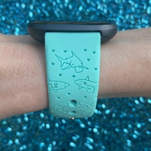 Pode incluir: Uma pulseira de rel&oacute;gio de silicone azul turquesa com um design de tubar&atilde;o e cora&ccedil;&atilde;o. A pulseira &eacute; compat&iacute;vel com os rel&oacute;gios inteligentes Fitbit.