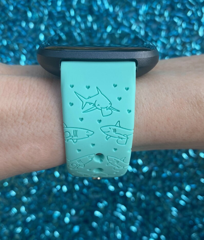 Pode incluir: Uma pulseira de rel&oacute;gio de silicone azul turquesa com um design de tubar&atilde;o e cora&ccedil;&atilde;o. A pulseira &eacute; compat&iacute;vel com os rel&oacute;gios inteligentes Fitbit.
