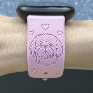 May include: A light pink watch band with a black watch face. The band features an engraved Shih Tzu dog design with hearts and paw prints. The band is attached to a wrist.