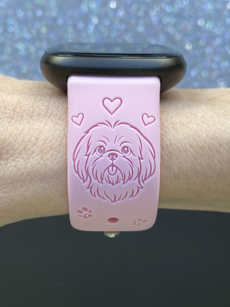 May include: A light pink watch band with a black watch face. The band features an engraved Shih Tzu dog design with hearts and paw prints. The band is attached to a wrist.