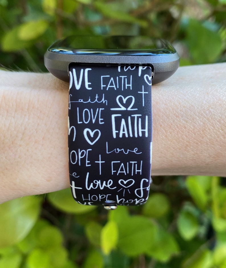 K&ouml;nnte beinhalten: Schwarz-wei&szlig;es Armband f&uuml;r eine Uhr mit den Worten "Love", "Faith" und "Hope", die sich in einem Muster wiederholen. Das Armband hat ein wei&szlig;es Herz- und Kreuzdesign.