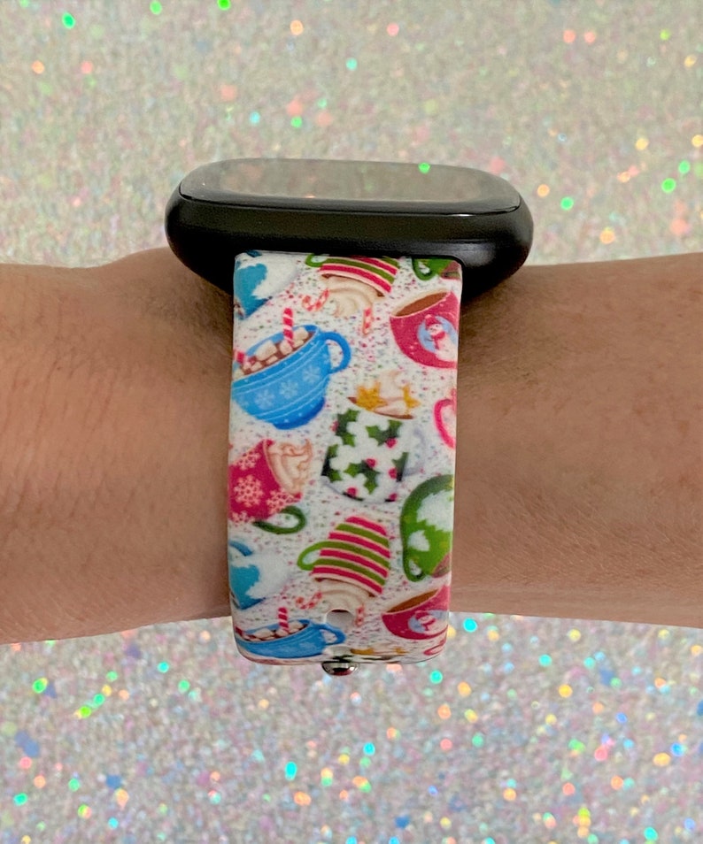 Puede incluir: Una correa de reloj blanca con un dise&ntilde;o navide&ntilde;o con tazas coloridas con chocolate caliente y bastones de caramelo.