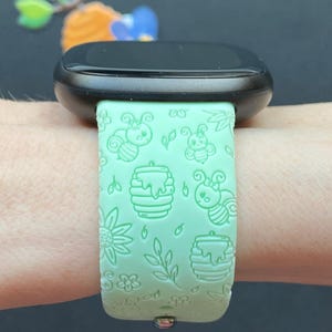 May include: A green silicone watch band with a bee and honeycomb pattern. The band is on a person's wrist.