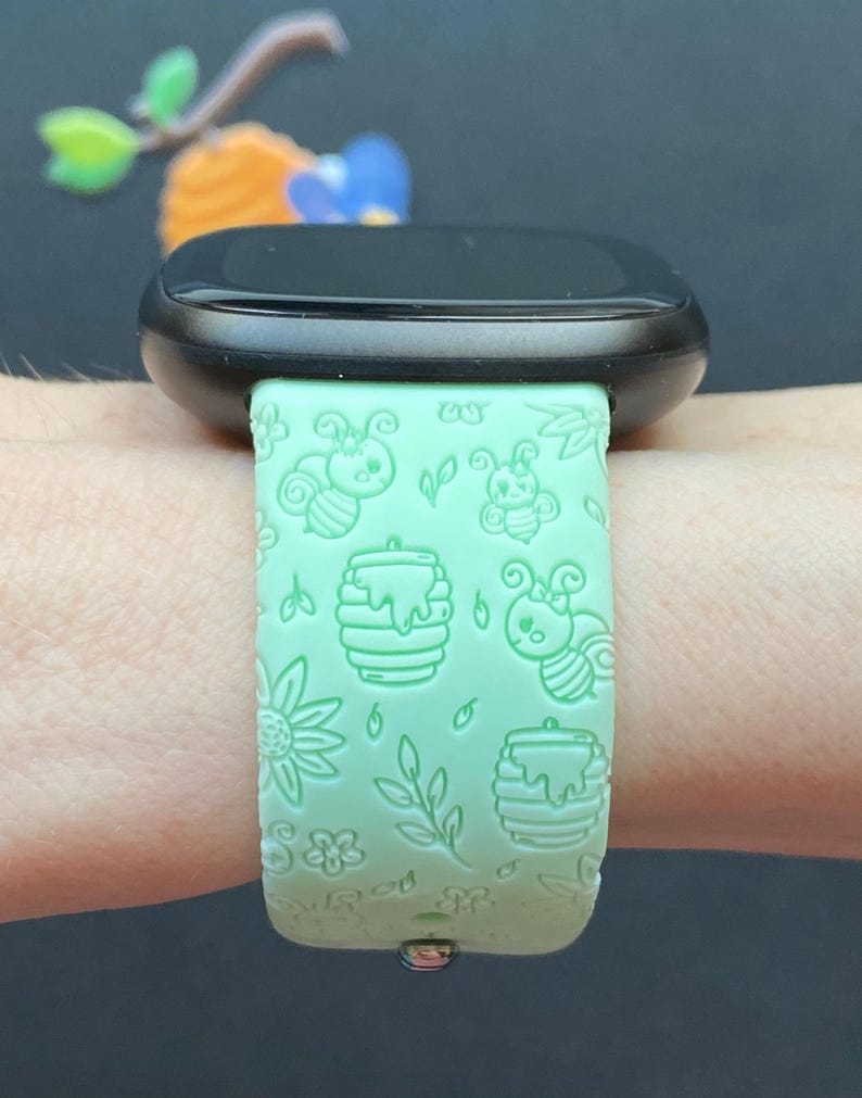 May include: A green silicone watch band with a bee and honeycomb pattern. The band is on a person's wrist.