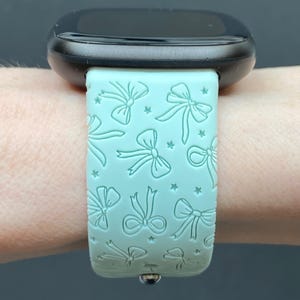 May include: A black smartwatch with a light green band featuring a repeating pattern of bows and stars. The band is made of a flexible material and is attached to the watch face with a silver clasp. The watch face is black and has a rounded shape.