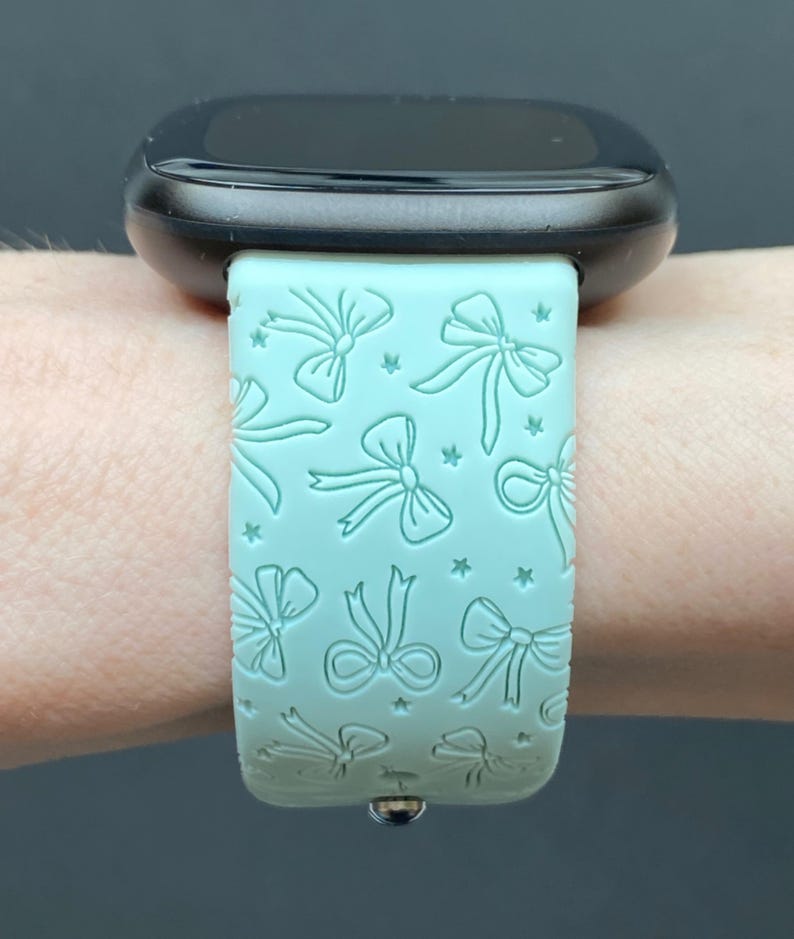 May include: A black smartwatch with a light green band featuring a repeating pattern of bows and stars. The band is made of a flexible material and is attached to the watch face with a silver clasp. The watch face is black and has a rounded shape.