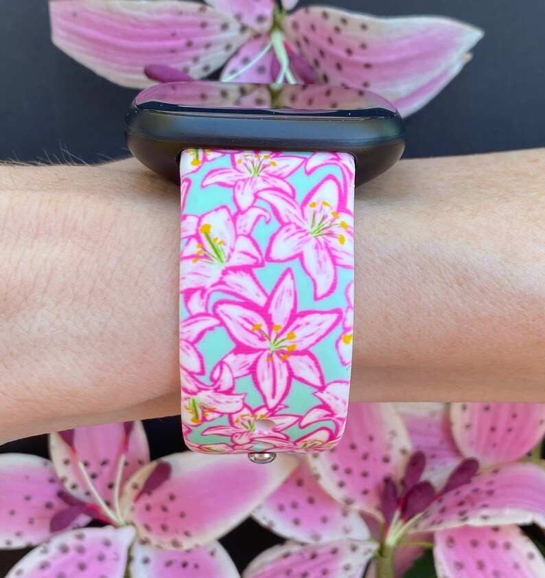 Puede incluir: Un reloj inteligente negro con una banda de patr&oacute;n floral rosa y verde. La banda presenta un patr&oacute;n repetido de lirios rosas sobre un fondo verde claro.