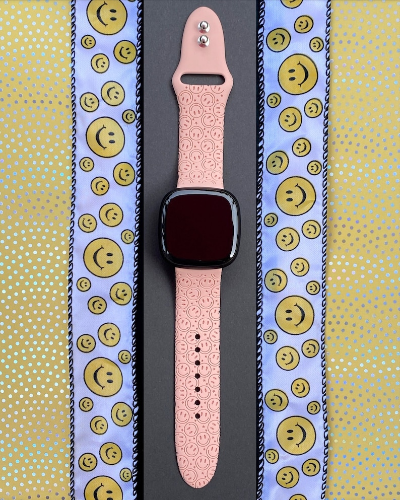 May include: A pink smartwatch band with a repeating pattern of smiley faces. The band is attached to a black smartwatch with a dark screen.