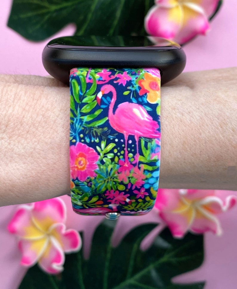 May include: A pink flamingo and tropical floral print watch band for a smartwatch. The band is made of silicone and has a navy blue background.