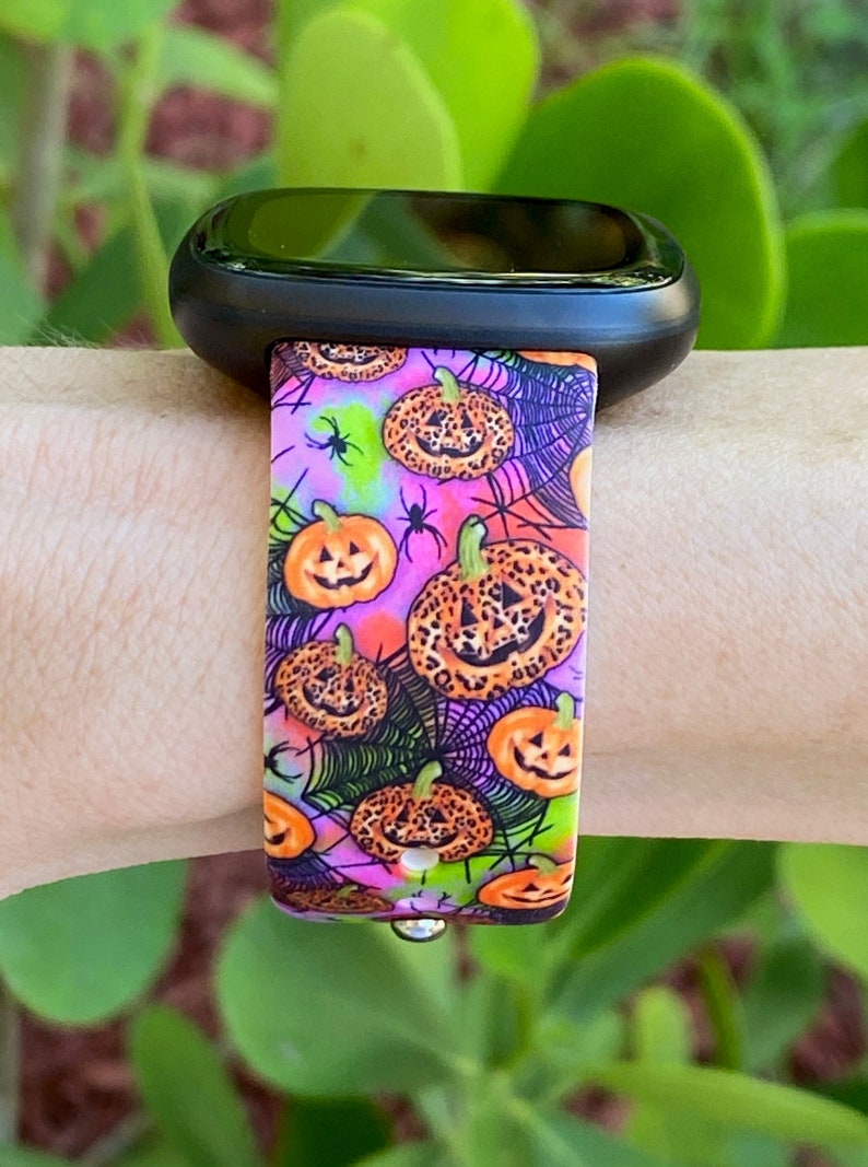 May include: A pink, black, green, and orange watch band with a pattern of pumpkins and spiderwebs. The pumpkins have a leopard print design.