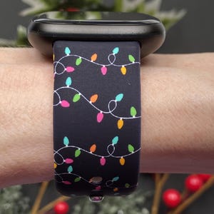 May include: Black smartwatch with a festive band featuring a string of colorful Christmas lights. The band has a dark background with white string and multi-colored bulbs. The watch is shown on a wrist.