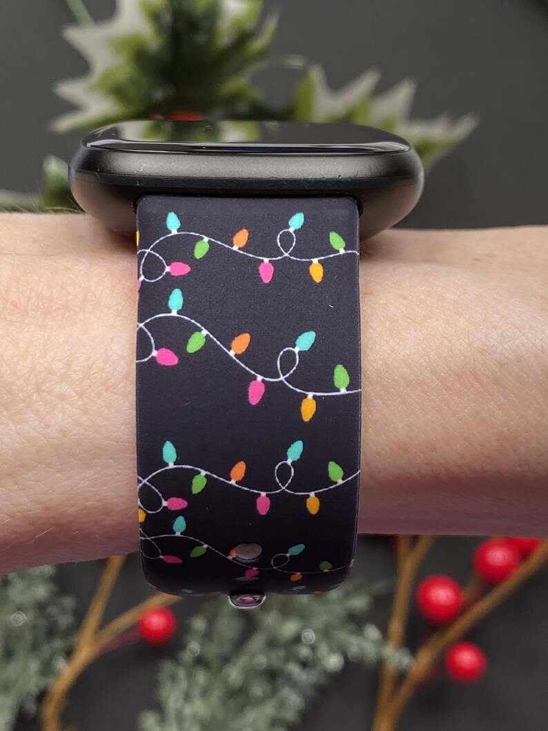 May include: Black smartwatch with a festive band featuring a string of colorful Christmas lights. The band has a dark background with white string and multi-colored bulbs. The watch is shown on a wrist.