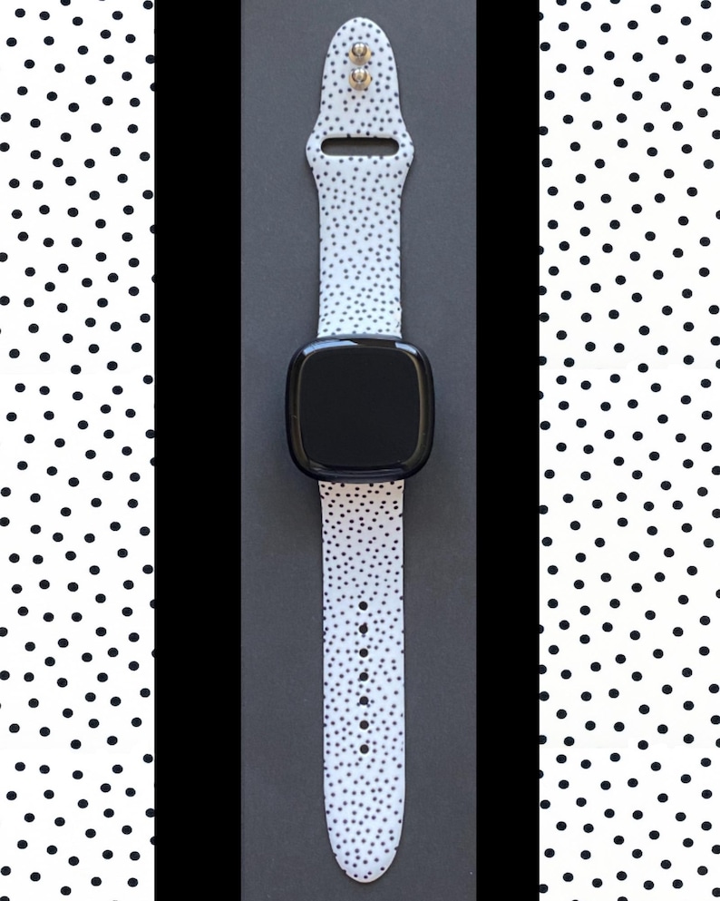 Pu&ograve; includere: Uno smartwatch nero con un cinturino bianco a pois neri. L'orologio ha uno schermo rettangolare e una cornice nera. Il cinturino ha fori di regolazione e una fibbia. Lo sfondo &egrave; una superficie nera.