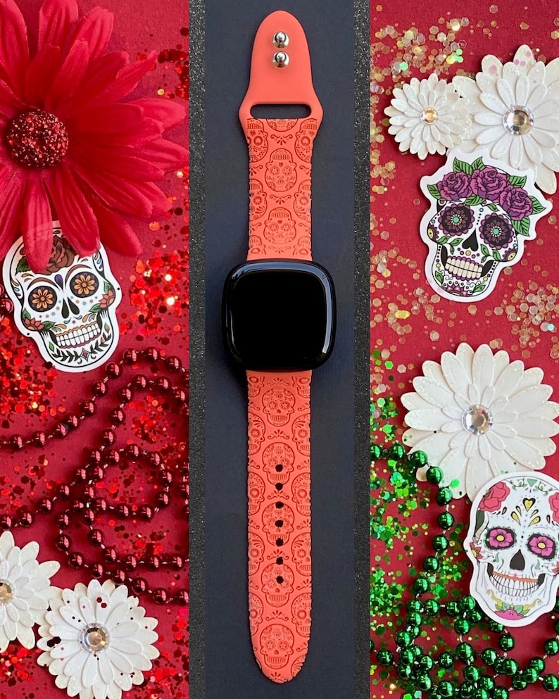 May include: A bright orange Apple Watch band with a repeating pattern of sugar skulls. The band is shown on a watch with a black face. The background is red with white flowers and green beads.