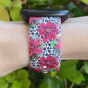 K&ouml;nnte beinhalten: Eine schwarze Smartwatch mit einem farbenfrohen Weihnachtsband. Das Band zeigt ein rotes Weihnachtsstern-Bl&uuml;tenmuster mit wei&szlig;en und schwarzen Leopardenmuster-Akzenten.