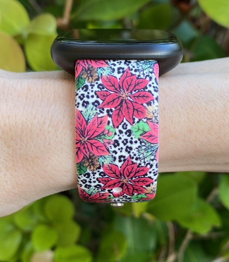 K&ouml;nnte beinhalten: Eine schwarze Smartwatch mit einem farbenfrohen Weihnachtsband. Das Band zeigt ein rotes Weihnachtsstern-Bl&uuml;tenmuster mit wei&szlig;en und schwarzen Leopardenmuster-Akzenten.