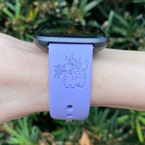 Peut inclure: Un bracelet de montre en silicone violet avec un motif de vache de dessin anim&eacute;. La vache tient un bouquet de fleurs dans sa bouche.