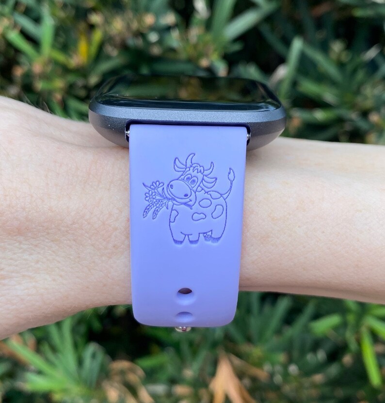 Peut inclure: Un bracelet de montre en silicone violet avec un motif de vache de dessin anim&eacute;. La vache tient un bouquet de fleurs dans sa bouche.