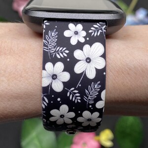 May include: Black smartwatch band with a white floral pattern. The band features a repeating design of large flowers and smaller leaves on a dark background. The watch face is black.