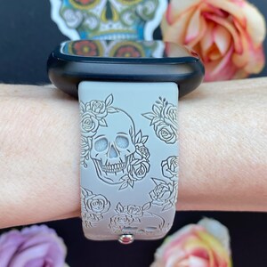 May include: A grey silicone watch band with a white skull and rose design. The band is on a person's wrist.