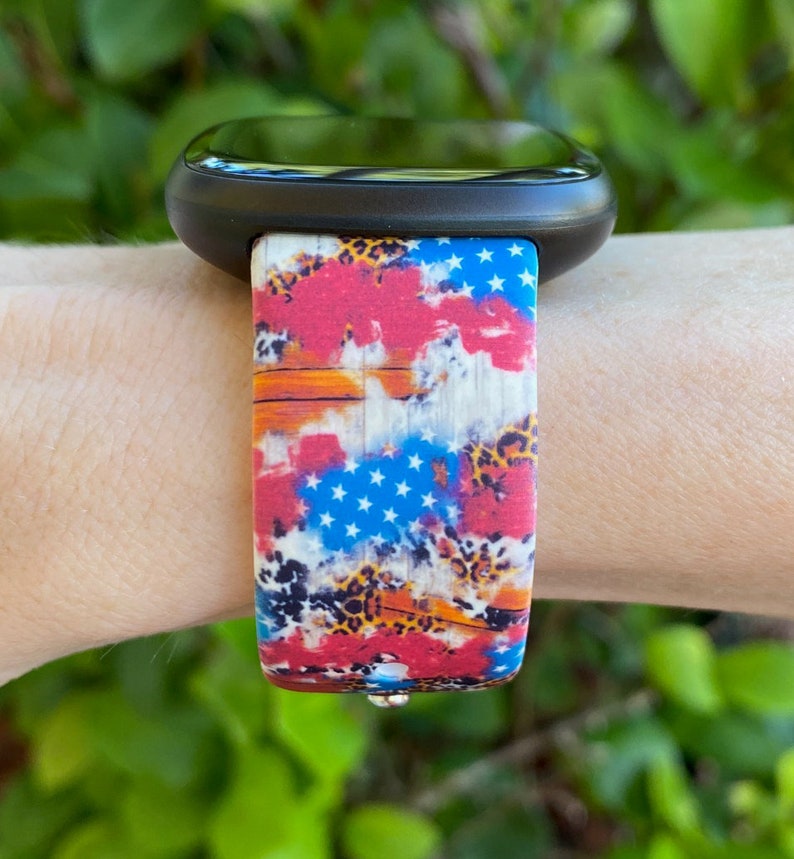 Pu&ograve; includere: Uno smartwatch nero con un cinturino colorato che presenta un design a bandiera americana rosso, bianco e blu con un motivo a stampa leopardata.
