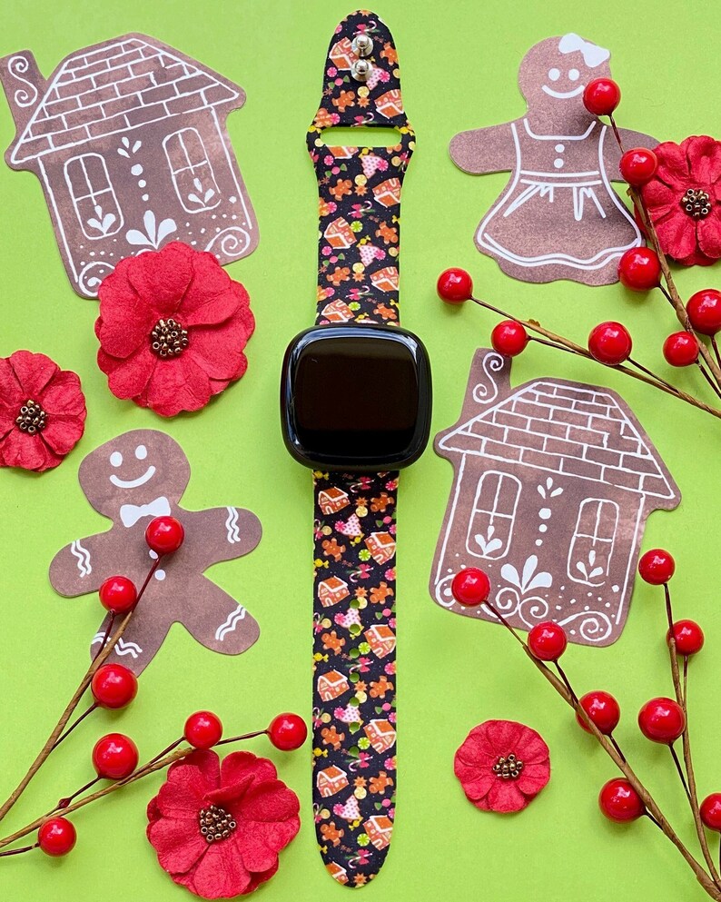 May include: A black smartwatch with a patterned band featuring gingerbread houses and cookies. The watch is surrounded by gingerbread cutouts, red flowers, and berry sprigs on a green background. A festive holiday theme.