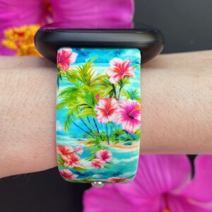 May include: A black smartwatch with a tropical floral print band. The band features pink flowers, palm trees, and a blue ocean scene.