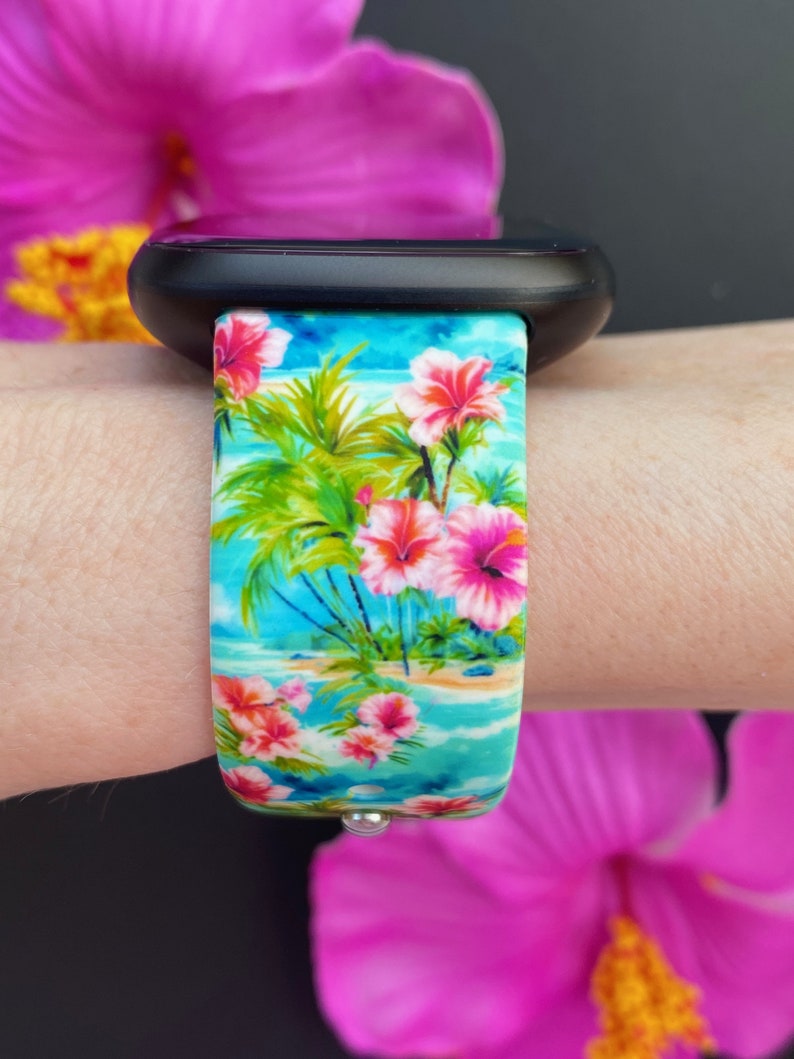 May include: A black smartwatch with a tropical floral print band. The band features pink flowers, palm trees, and a blue ocean scene.
