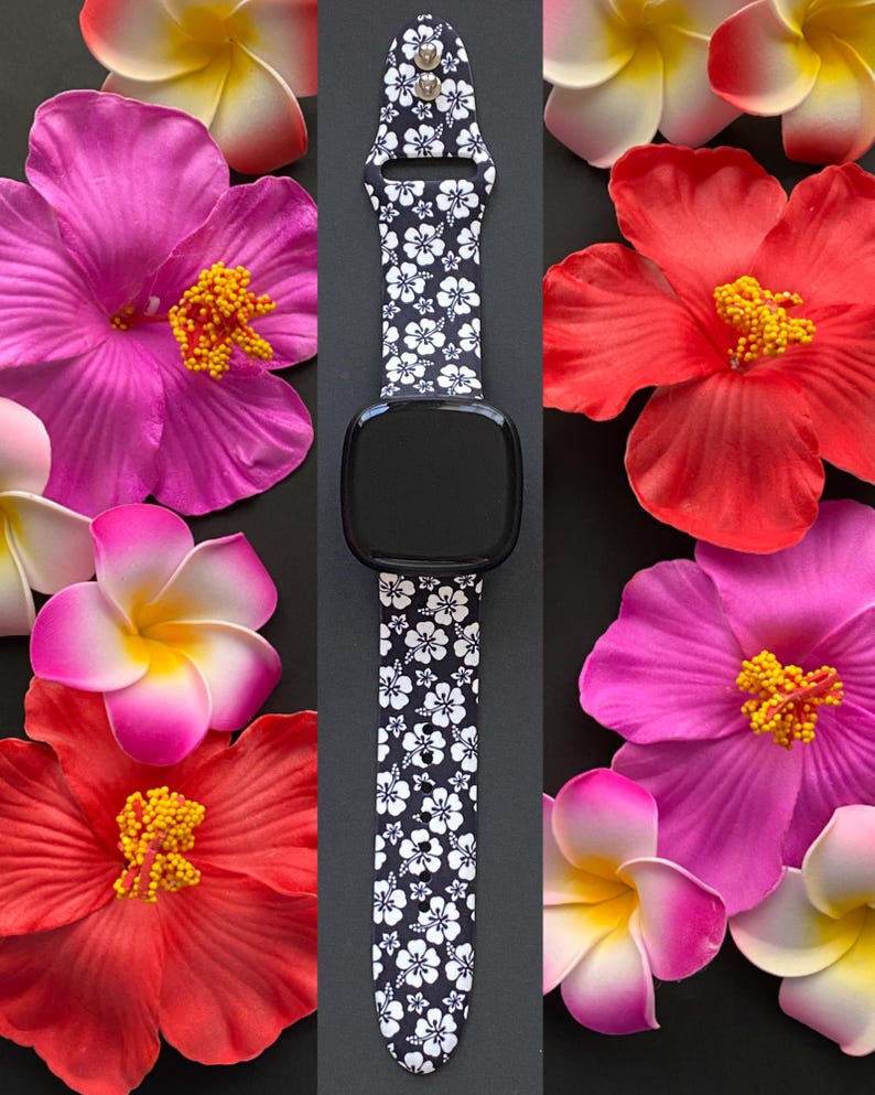 May include: Black smartwatch with a white floral pattern band. The watch is surrounded by vibrant pink, red, and yellow artificial flowers on a black background. The watch band features a repeating white flower design.