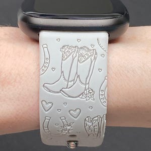 May include: White silicone watch band with a design of cowboy boots, hearts, and horseshoes. The band is compatible with a smartwatch.