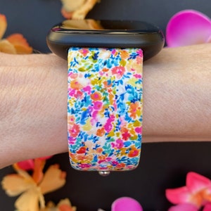 K&ouml;nnte beinhalten: Eine schwarze Smartwatch mit einem wei&szlig;en Armband mit Blumenmuster. Das Armband hat verschiedene Farben, darunter Pink, Blau, Gelb und Gr&uuml;n.