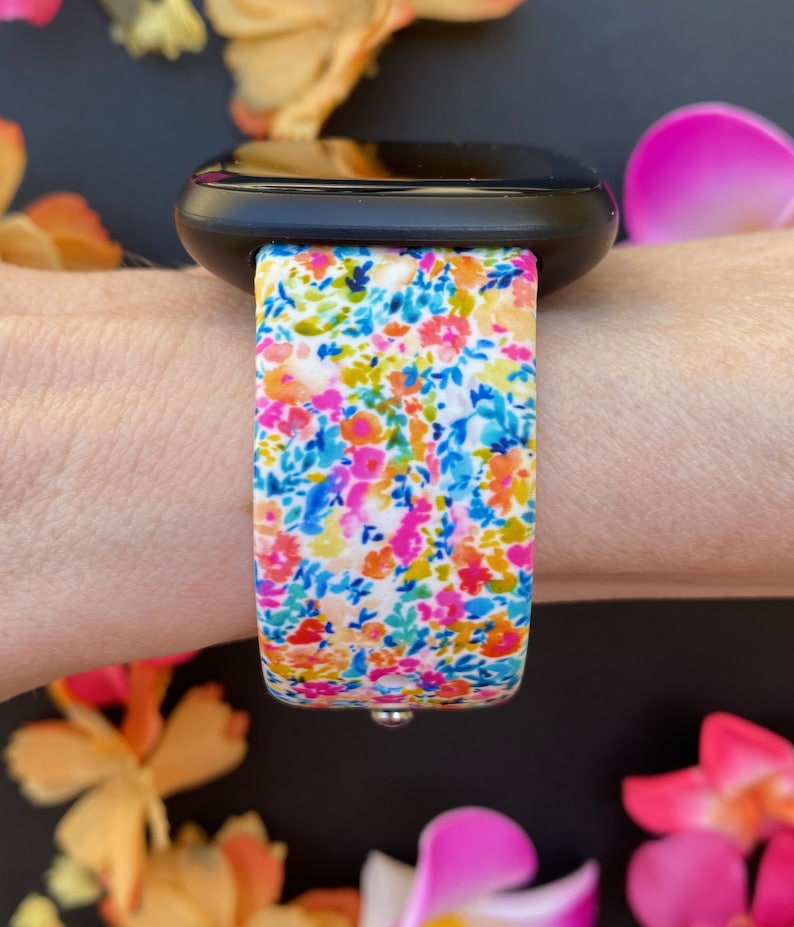 K&ouml;nnte beinhalten: Eine schwarze Smartwatch mit einem wei&szlig;en Armband mit Blumenmuster. Das Armband hat verschiedene Farben, darunter Pink, Blau, Gelb und Gr&uuml;n.