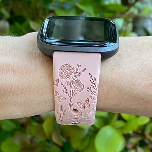 May include: A pink silicone watch band with a floral and butterfly design. The band is compatible with a Fitbit watch.
